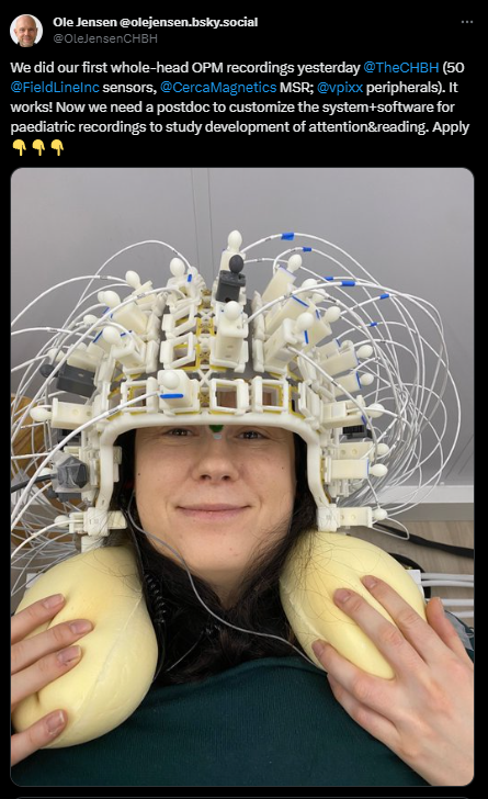 A screenshot of a social media post from Dr. Ole Jensen, with the text" "We did our first whole-head OPM recordings yesterday 
@TheCHBH
 (50 
@FieldLineInc
 sensors, 
@CercaMagnetics
 MSR; 
@vpixx
 peripherals). It works! Now we need a postdoc to customize the system+software for paediatric recordings to study development of attention&reading. Apply below"

Below text is an image of a woman smiling at the camera. She is wearing a white helmet with sensors and cabling emanating from it in a halo. She is also wearing a beige neck cushion for support.